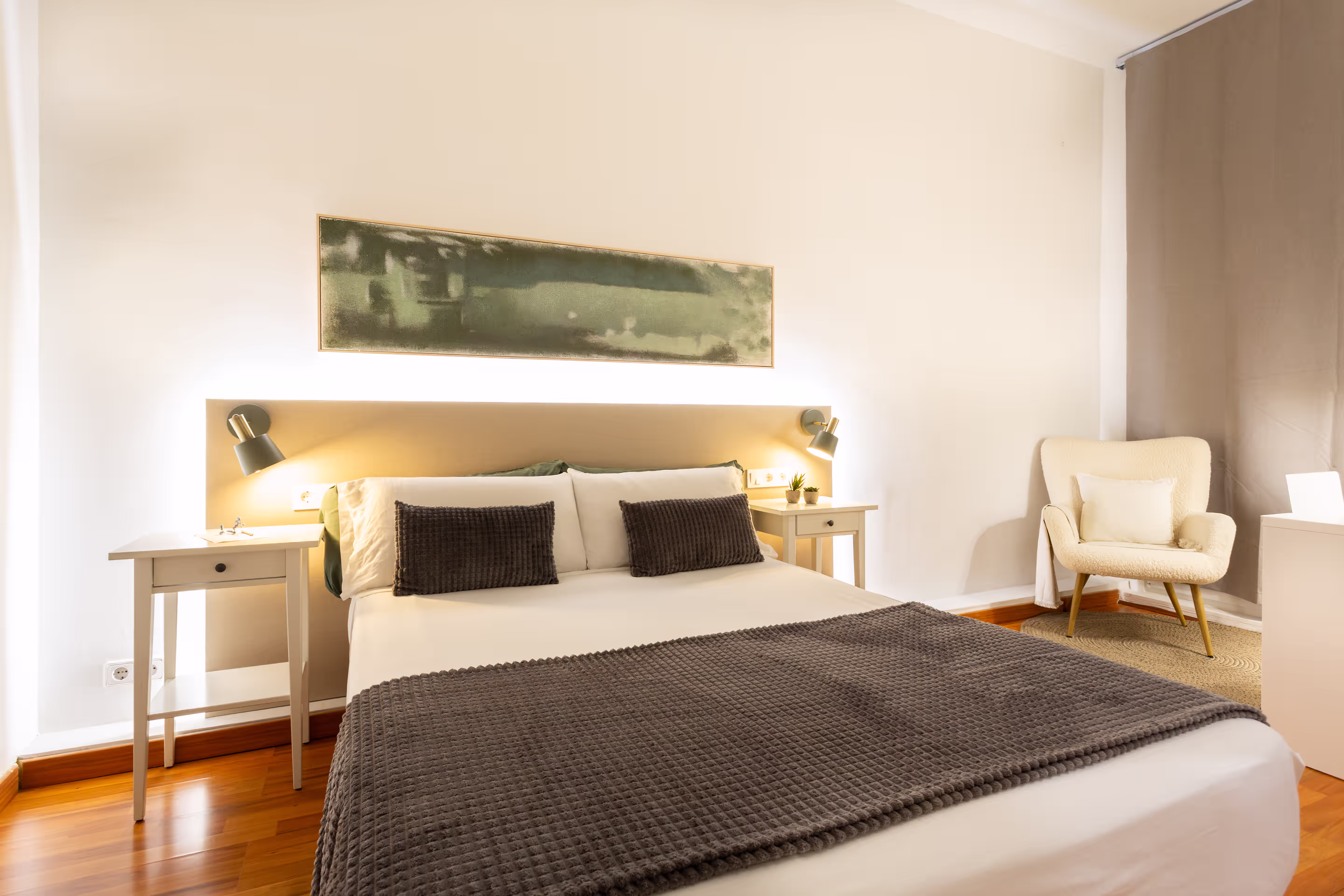 Minimalist bedroom with a bed covered in white and gray linens, two side tables, and a beige armchair.