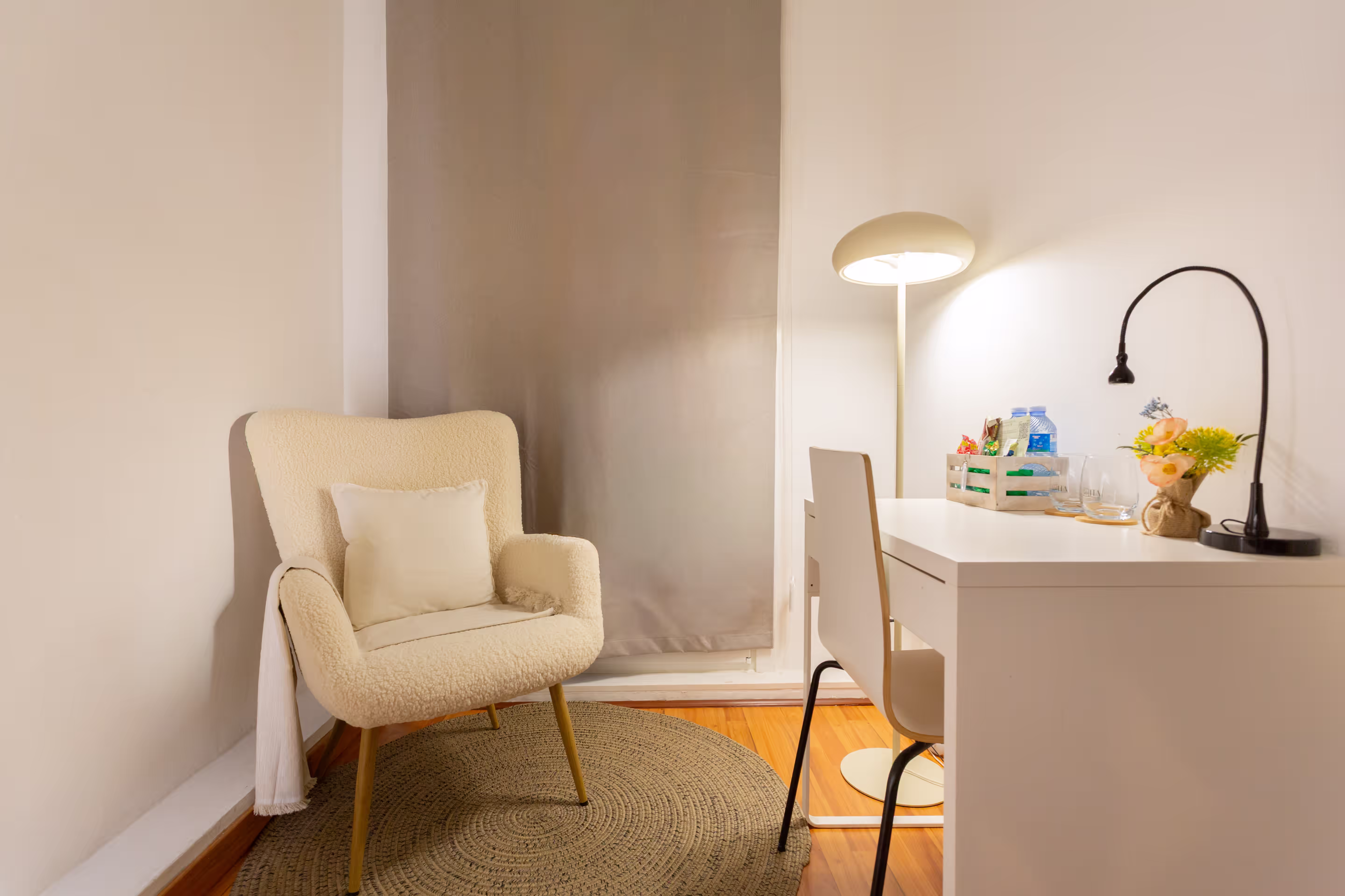 Cozy cream armchair with pillow on circular rug beside a white desk with lamps and flower vase.