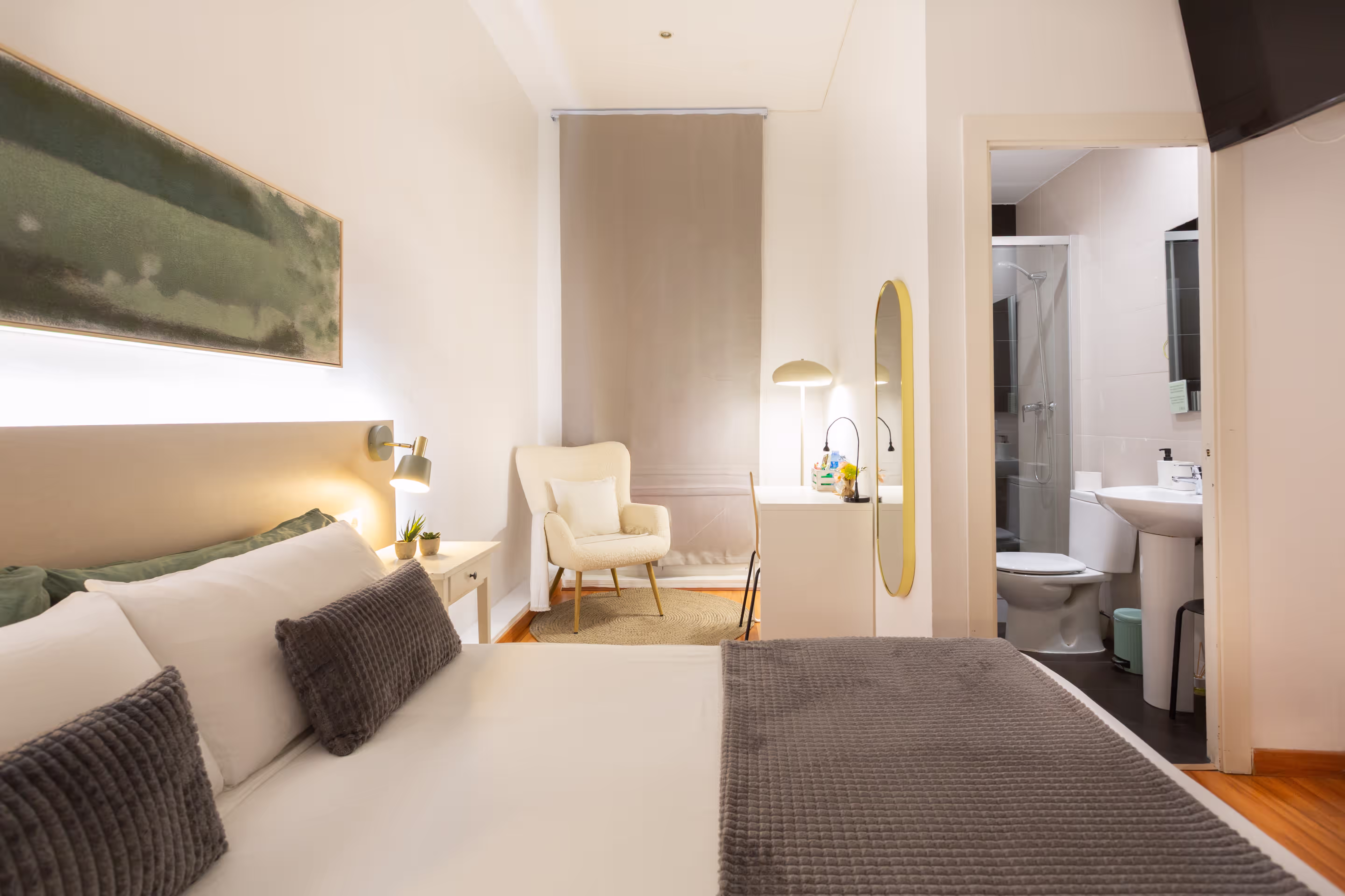 Bright bedroom with white bed, gray pillows, armchair, white desk, and visible bathroom door.