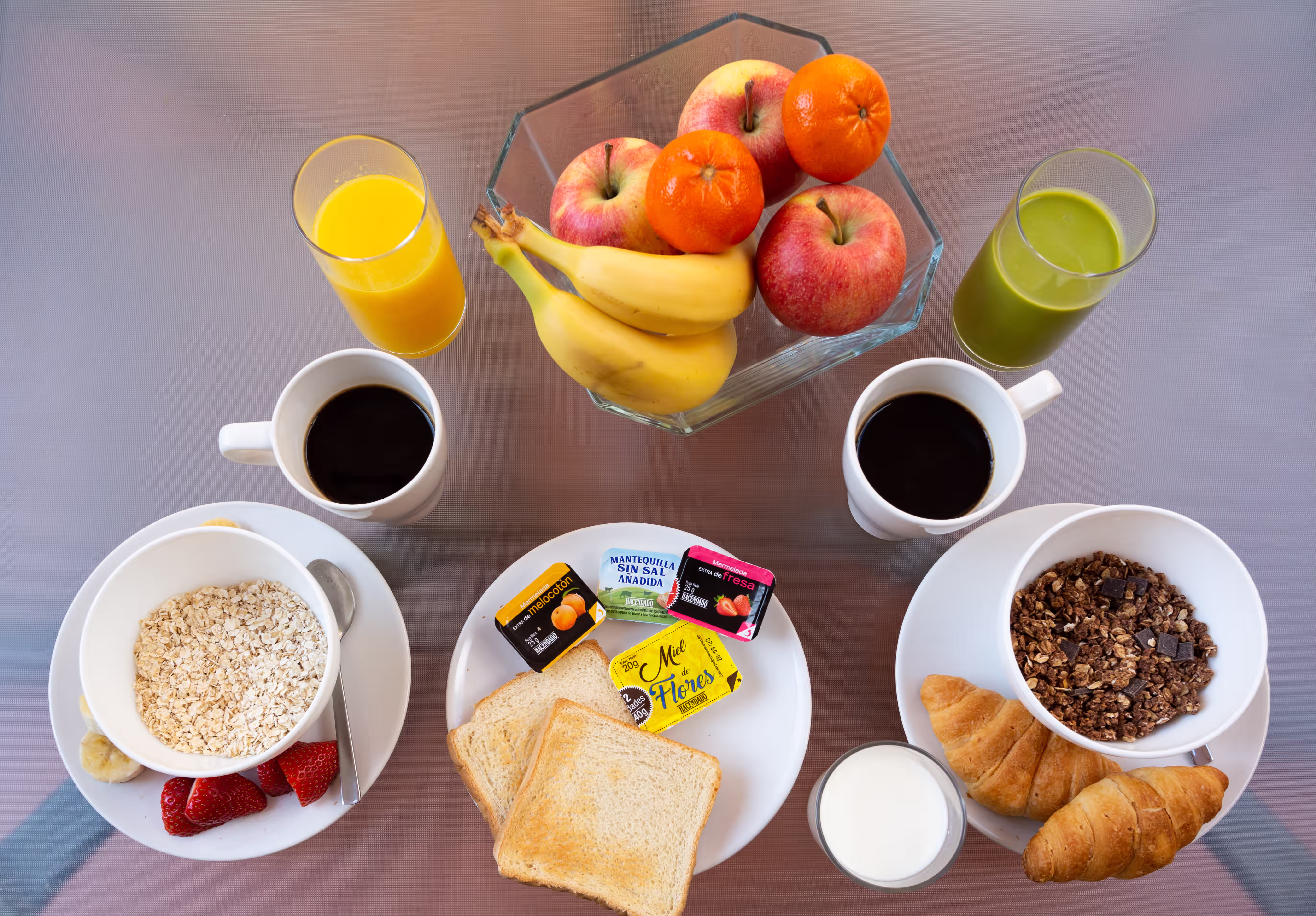 Breakfast spread with oats, strawberries, bananas, toast, croissants, coffee, milk, juices, and a fruit bowl.