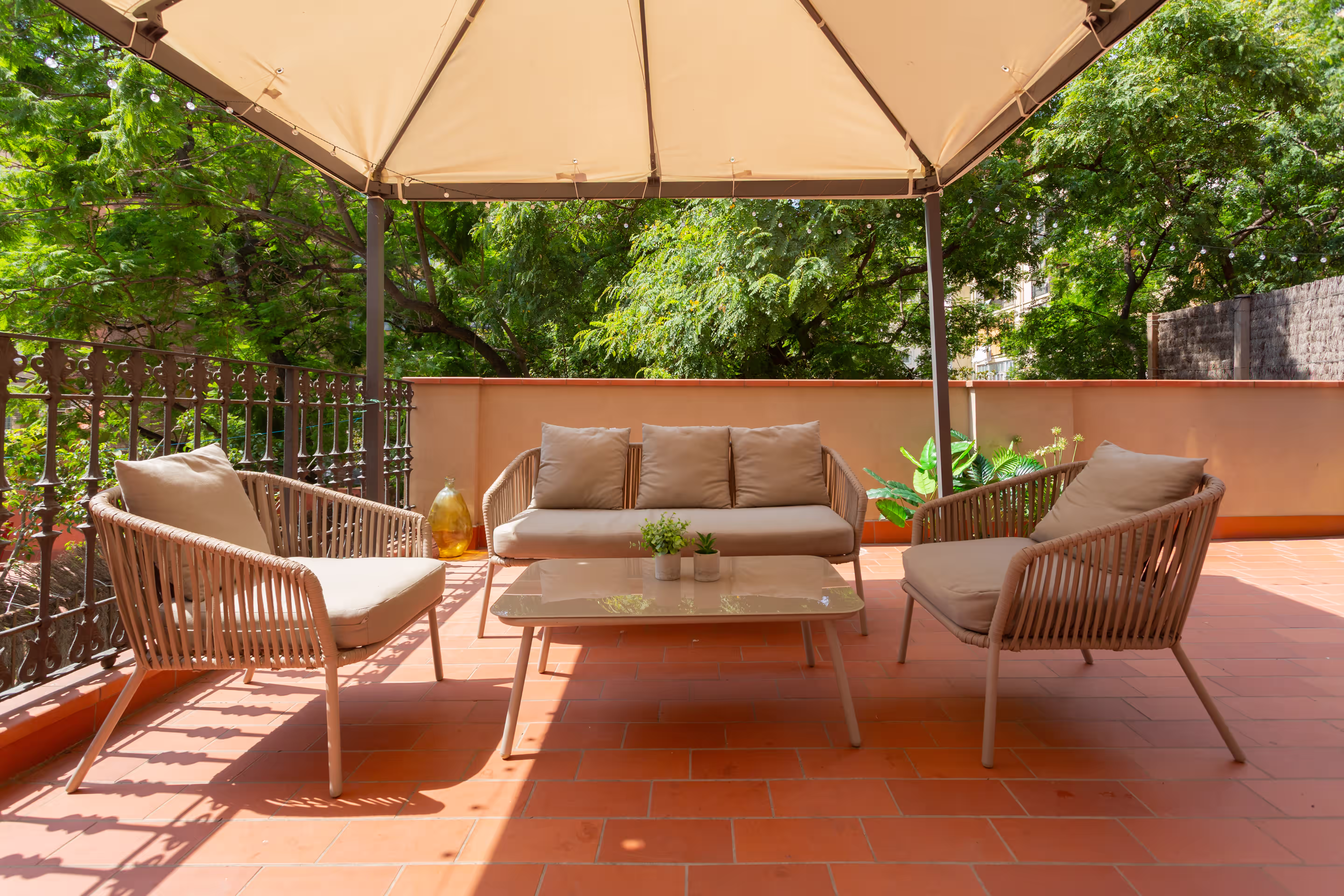 Outdoor patio seating with beige cushioned chairs, sofa, table, plants, and a tan canopy overhead.