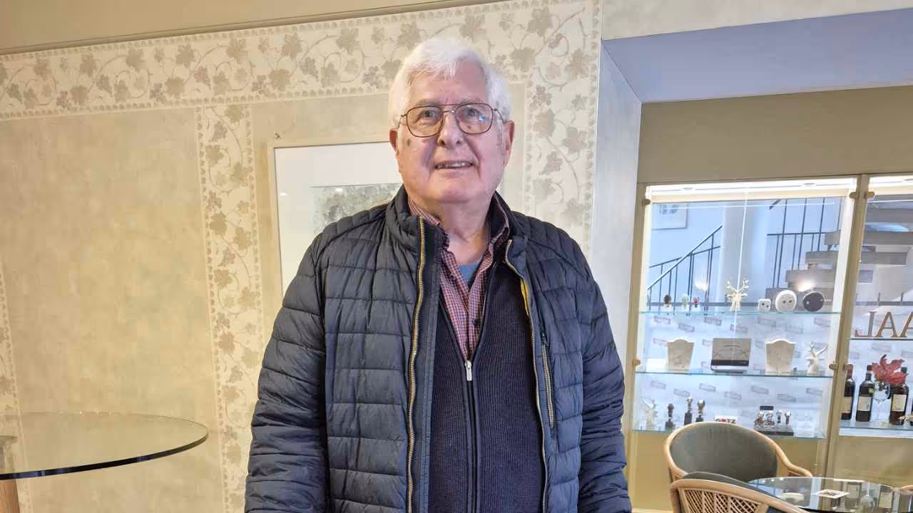 Porträt eines Mannes mit weißen Haaren und Brille in einer hellen Inneneinrichtung mit Vitrine und Weinlaub-Tapete.