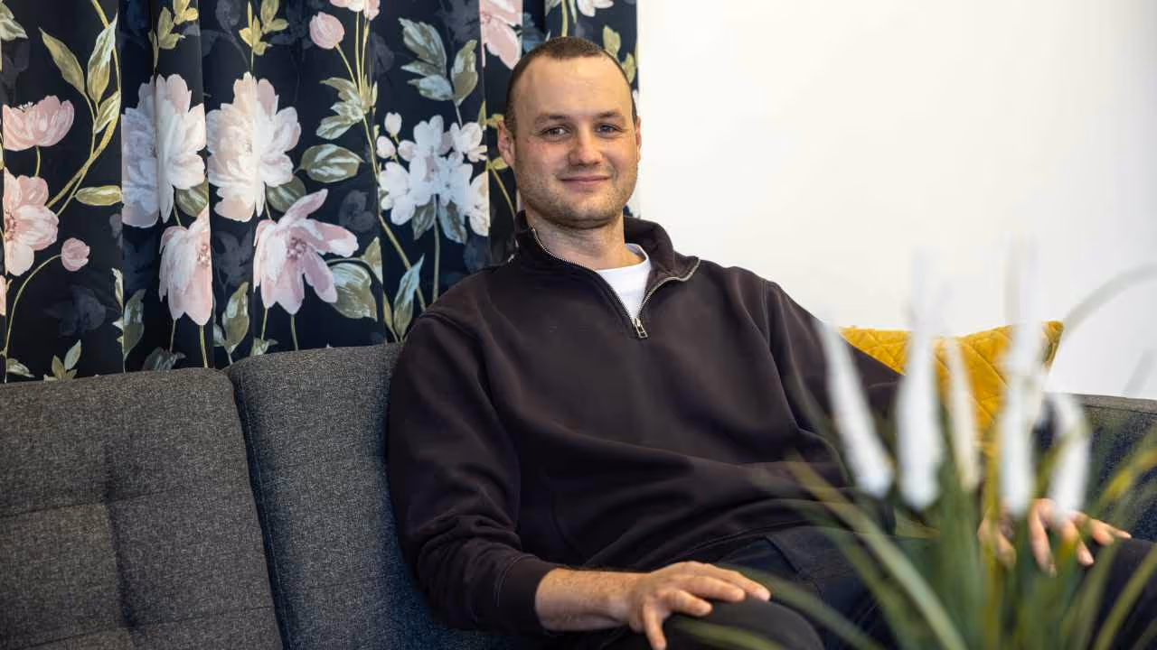 Porträt eines Mannes im dunklen Pullover auf einer Couch, im Hintergrund ein floraler Vorhang und helle Wand.