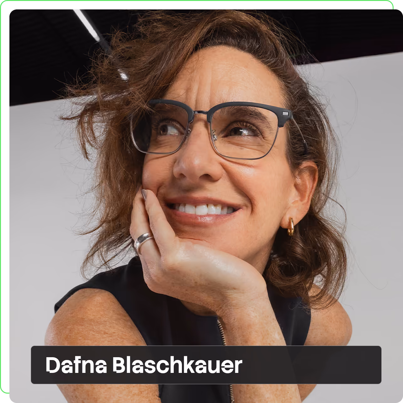 Dafna Blaschkauer sorrindo com a mão apoiada no rosto, cabelo castanho ondulado, óculos de armação escura e blusa preta sem mangas, em estúdio de fundo claro