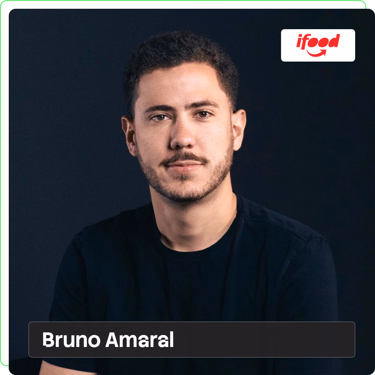 Bruno Amaral olhando para a câmera com leve sorriso, cabelo curto escuro, barba baixa e camiseta preta, retrato em estúdio com fundo azul-escuro