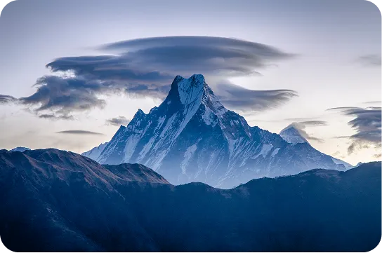 Snow-covered mountain peak with dramatic cloud formations in a clear sky at dusk.