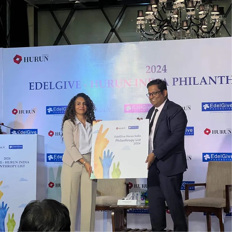 Two people holding a large plaque that reads 'EdelGive Hurun India Philanthropy List 2024' at an event with branded backdrop and chandelier.