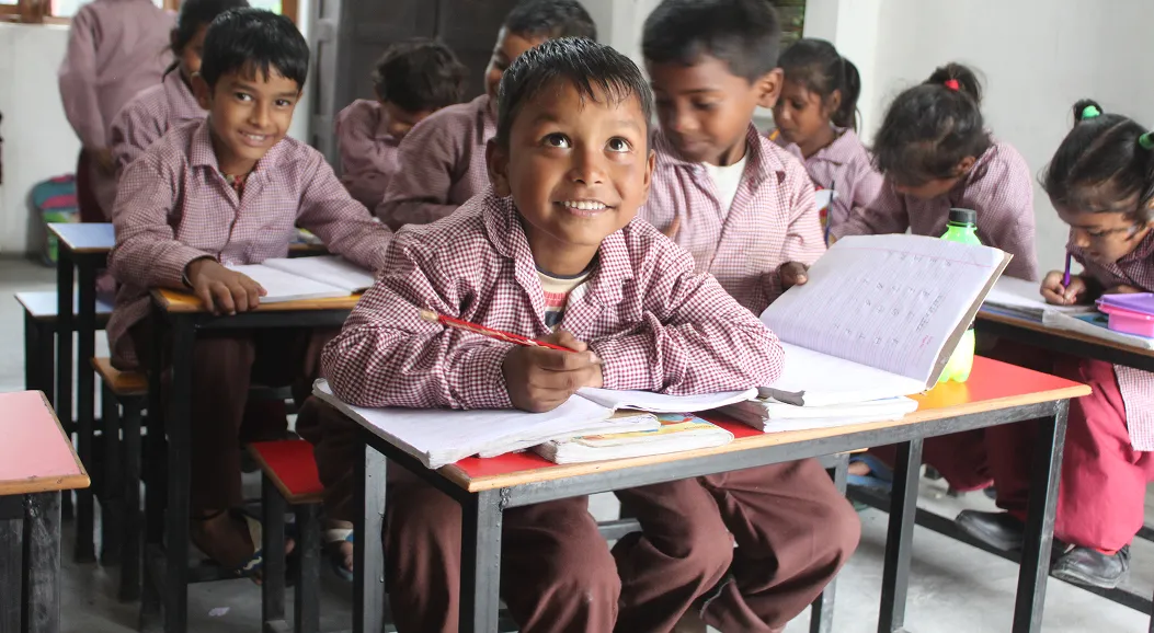 A group of young children in matching checkered school uniforms sitting at desks in a classroom, with one boy in the foreground smiling and holding a pencil over an open notebook.