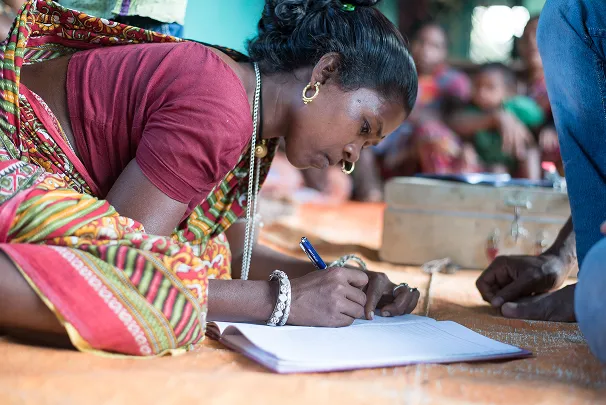 Woman in traditional colorful attire writing in a notebook while sitting on the floor indoors.