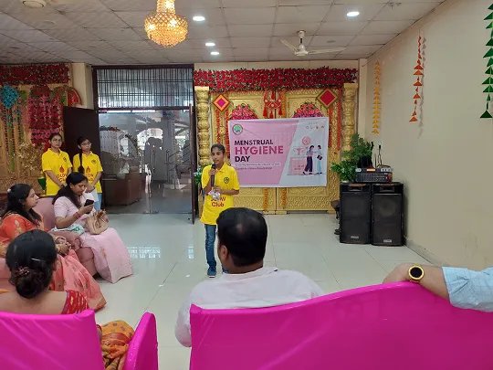 A young girl in a yellow shirt speaking into a microphone in a decorated room with a Menstrual Hygiene Day banner, while an audience watches.