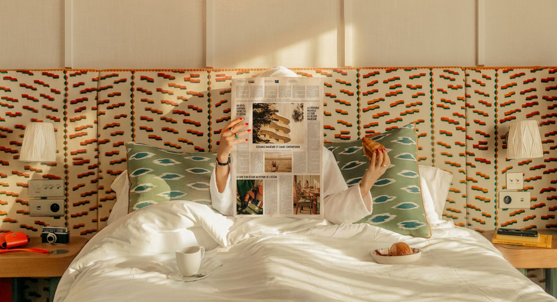Personne au lit avec peignoir blanc, lisant un journal et tenant un croissant, entourée de coussins verts et décor rétro.