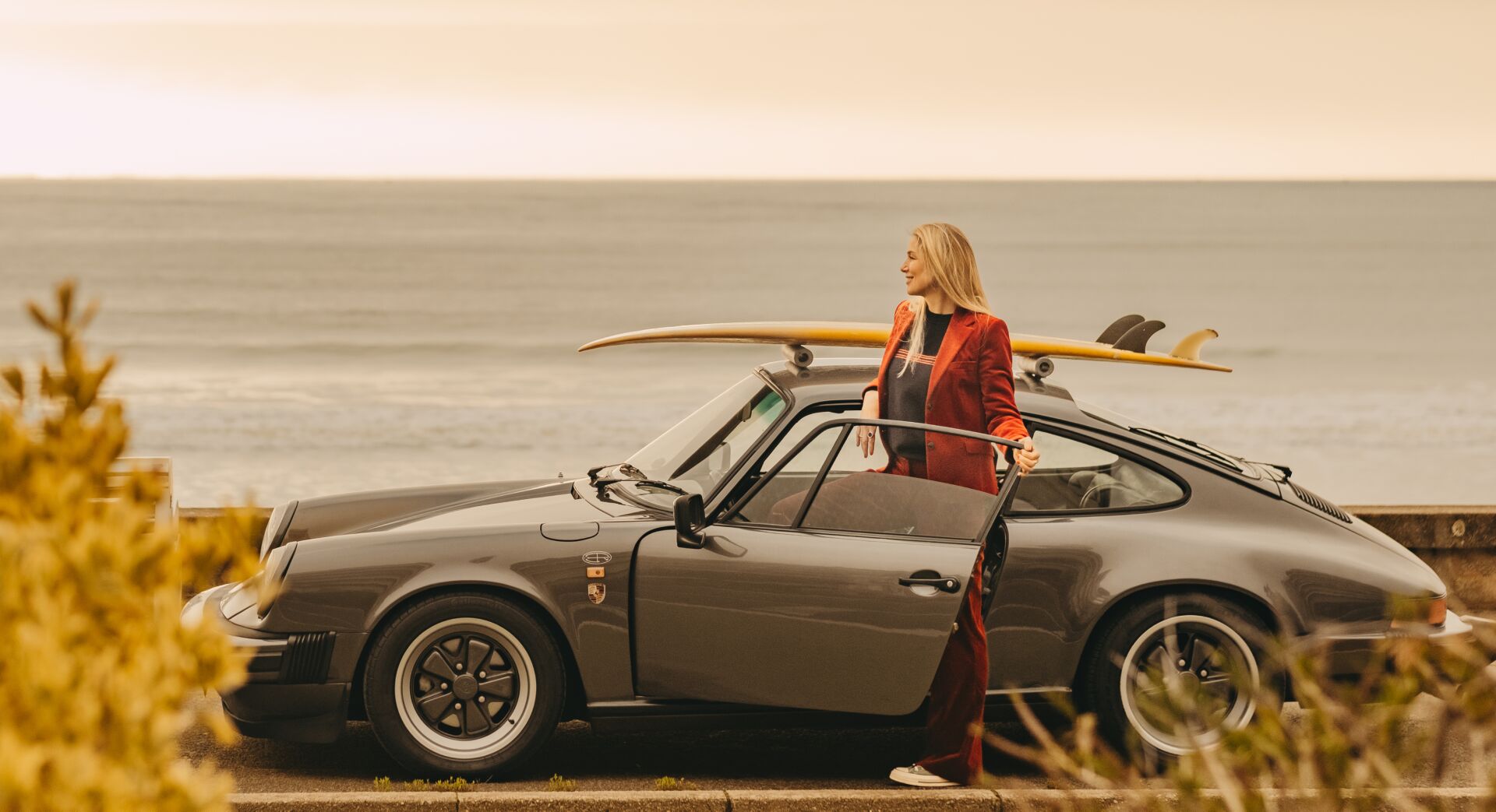 Femme vêtue d'un tailleur rouge ouvrant la porte d'une voiture de sport grise avec une planche de surf jaune sur le toit au bord de la mer.