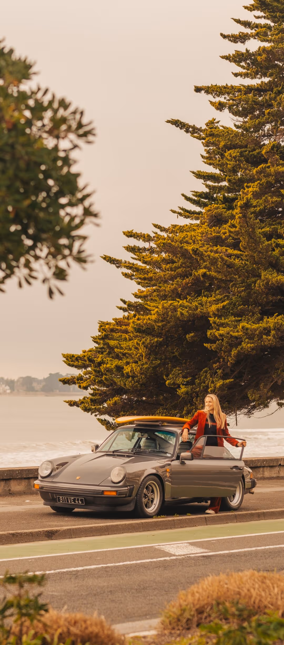 Une femme debout près d’une voiture classique grise avec une planche de surf sur le toit, stationnée en bord de mer sous de grands arbres.