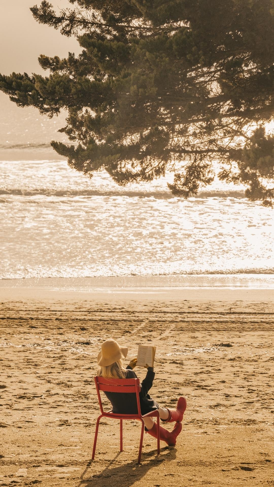 Une personne assise sur une chaise rouge lit un livre sur la plage, sous un grand pin, face à la mer.