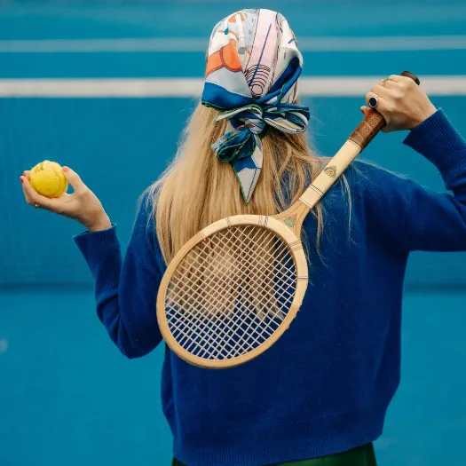 Joueuse de tennis élégante avec raquette en bois et foulard coloré sur court bleu, art de vivre baulois aux Cimes Bleues.