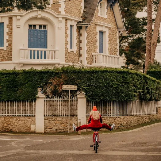 Femme en tenue rouge à vélo devant une villa balnéaire typique de La Baule, balade légère et joyeuse à découvrir depuis les Cimes Bleues.