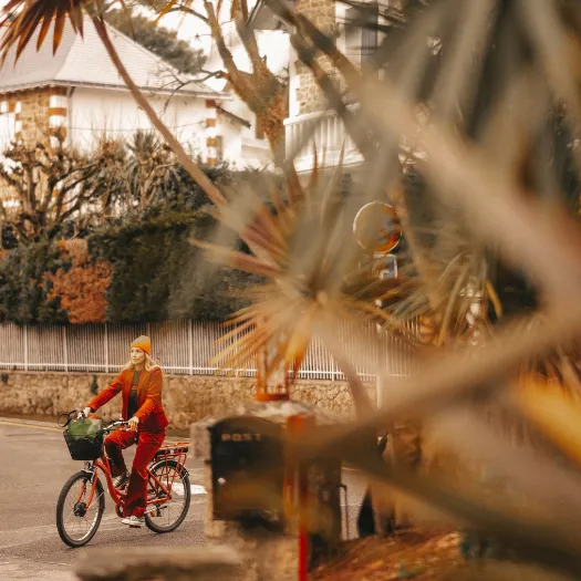 Femme à vélo en tenue terracotta dans les ruelles boisées de La Baule, balade nature au départ des Cimes Bleues, hôtel 4 étoiles.