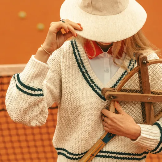 Elegant tennis player in a cream tennis sweater and beige hat on a clay court, with a sport chic vibe from Cimes Bleues in La Baule.