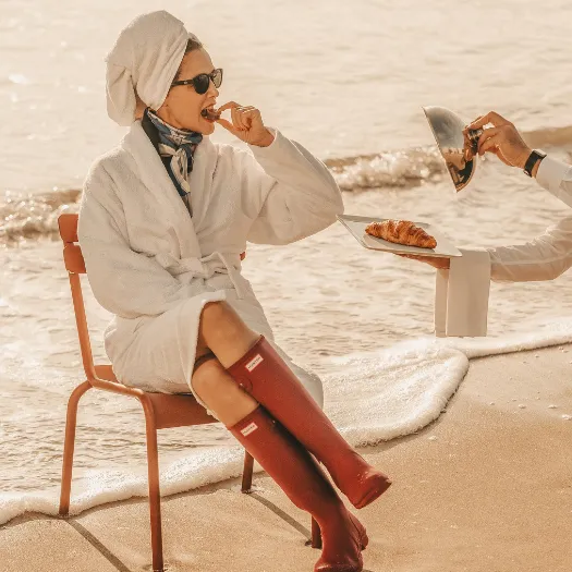 Woman in a white bathrobe enjoying a croissant with her feet in the water on La Baule beach, room service and the art of living at Les Cimes Bleues.