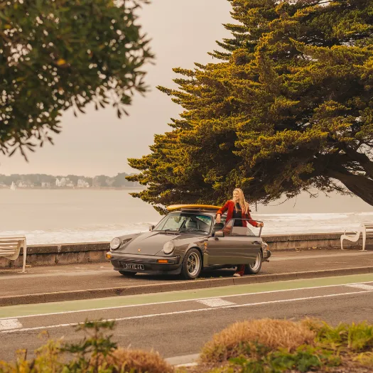 A woman in a red jacket leaning on a vintage Porsche 911 convertible on the La Baule seafront, a picture of elegance and freedom, embodying the spirit of Cimes Bleues.