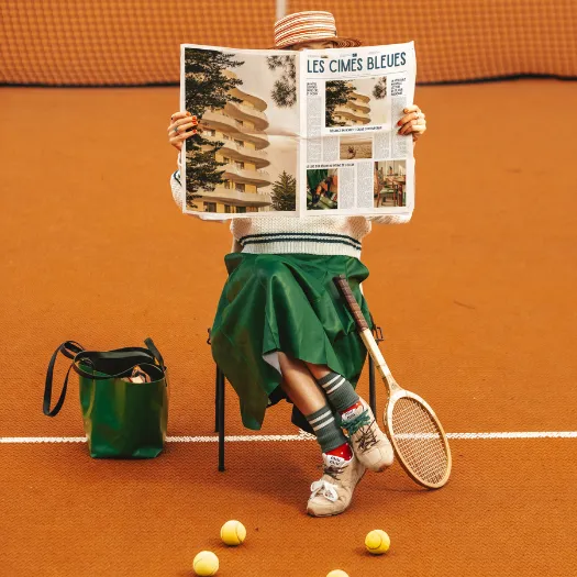 Tennis player reading the Les Cimes Bleues newspaper on a clay court, embodying the sporting spirit and sophisticated lifestyle of the 4-star hotel in La Baule.