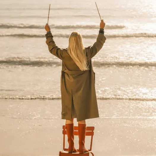 Woman in a trench coat and red boots, commanding the ocean view as she faces the waves of La Baule, embodying freedom and unconventional elegance in the spirit of Cimes Bleues.