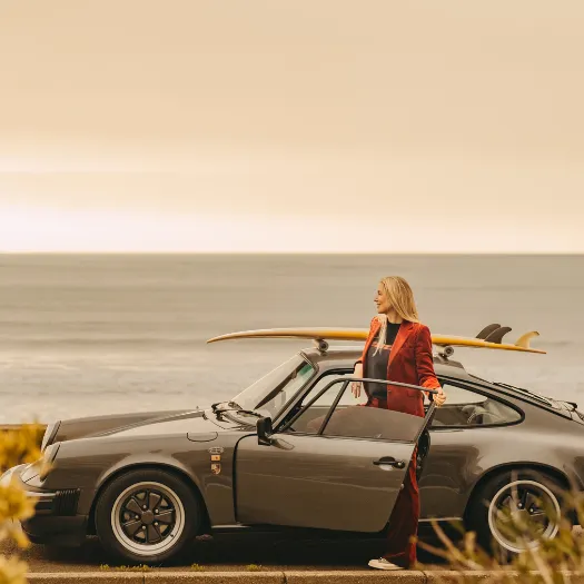 Woman in a red jacket stepping out of a vintage Porsche 911 with a surfboard on the roof, facing the ocean in La Baule, freedom and elegance at Les Cimes Bleues.