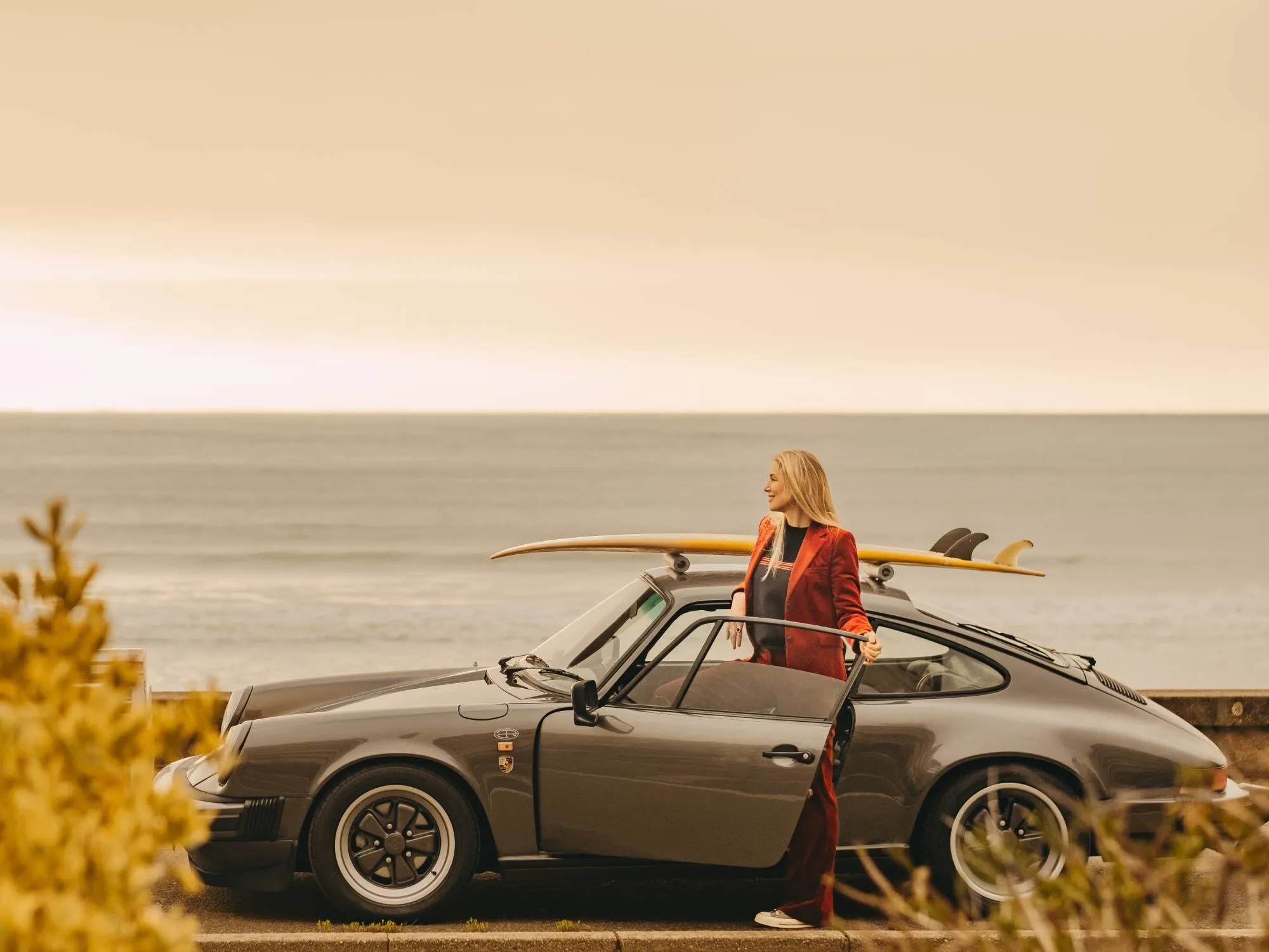 A blonde woman getting into her Porsche with a surfboard on the roof and the sea in the background. 