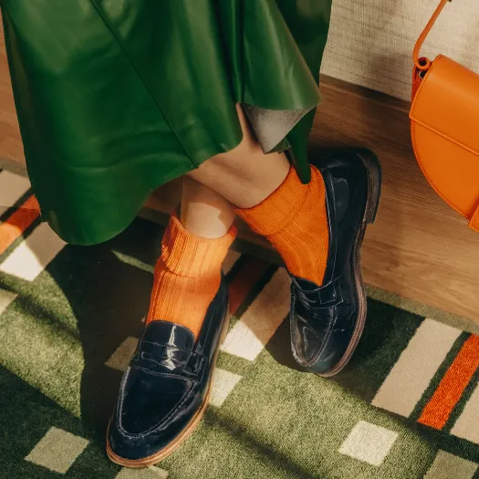 Fashion detail at Les Cimes Bleues: navy patent loafers, bright orange socks, and a green skirt on a graphic rug, a chic and colorful style in La Baule.