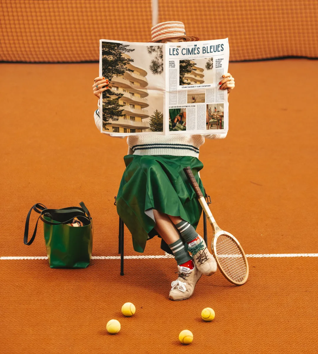 Joueuse de tennis assise sur court en terre battue lisant le journal Les Cimes Bleues, jupe verte et raquette en bois, esprit sport chic de l'hôtel 4 étoiles à La Baule.