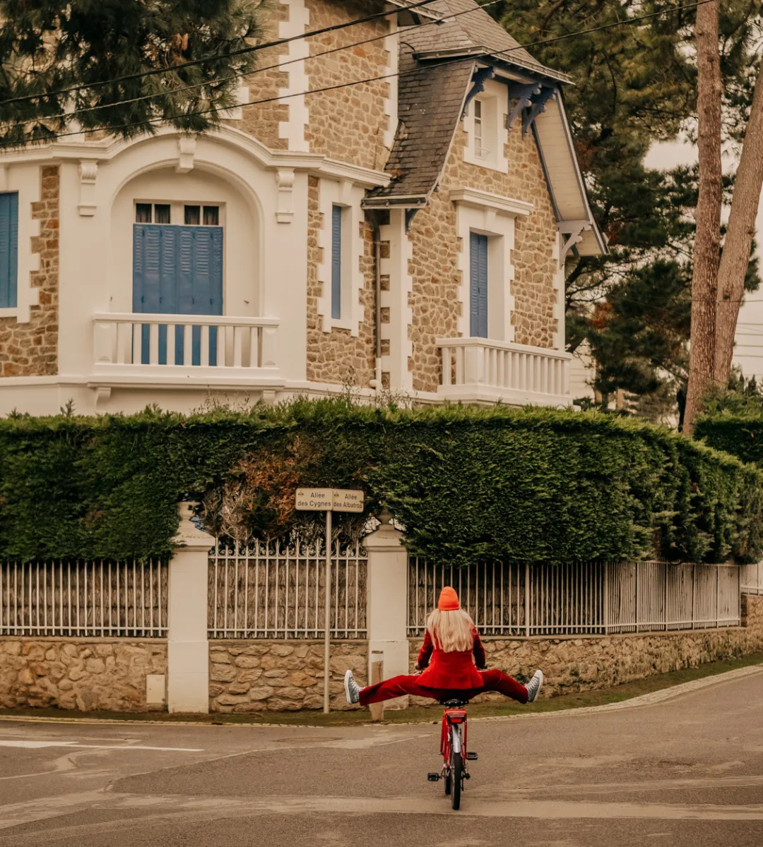 Femme en tenue rouge les bras en croix à vélo devant une villa balnéaire typique de La Baule, insouciance et joie de vivre dans l'esprit des Cimes Bleues.