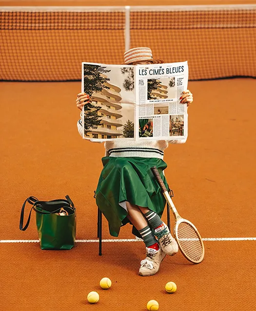Femme assise sur un court de tennis en terre battue lisant un journal, jupe verte et chapeau rayé, raquette vintage et balles de tennis au sol.