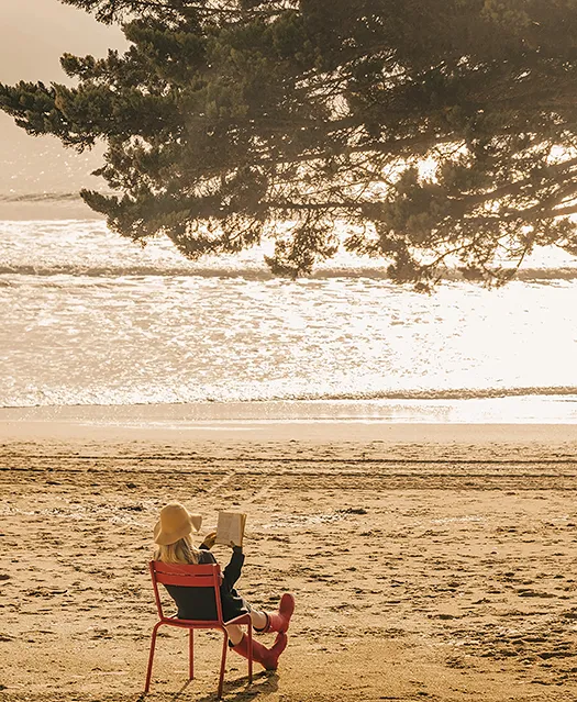 Femme assise sur une chaise rouge lisant un livre face à l'océan sur une plage de sable doré sous un pin maritime au coucher du soleil.