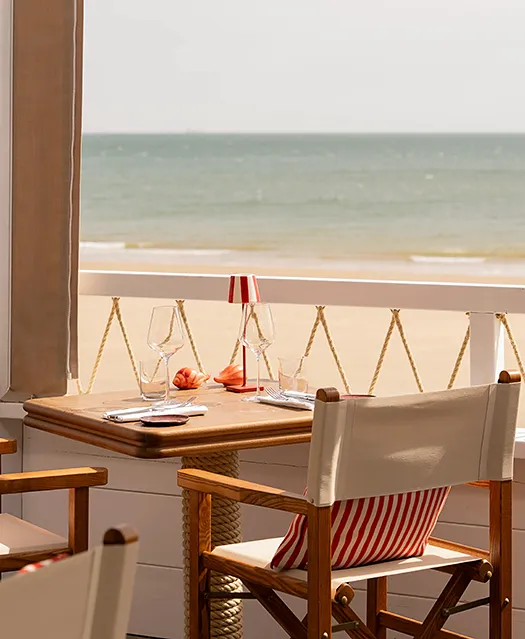 Table de restaurant en bord de mer dressée avec verres à vin et petite lampe rouge rayée sur terrasse avec vue.