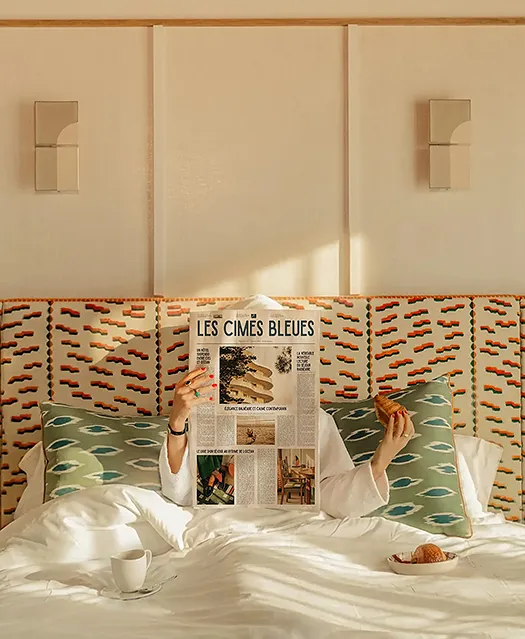 Femme dans un lit lisant un journal, un croissant à la main.