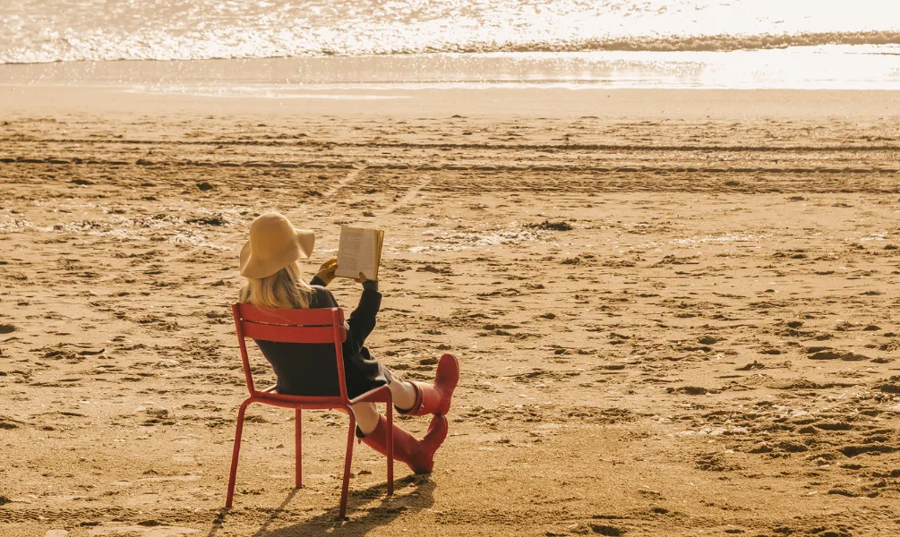 Femme au chapeau beige et bottes rouges installée sur une chaise Fermob en bord de mer avec vue sur les vagues et lumière dorée de fin de journée.