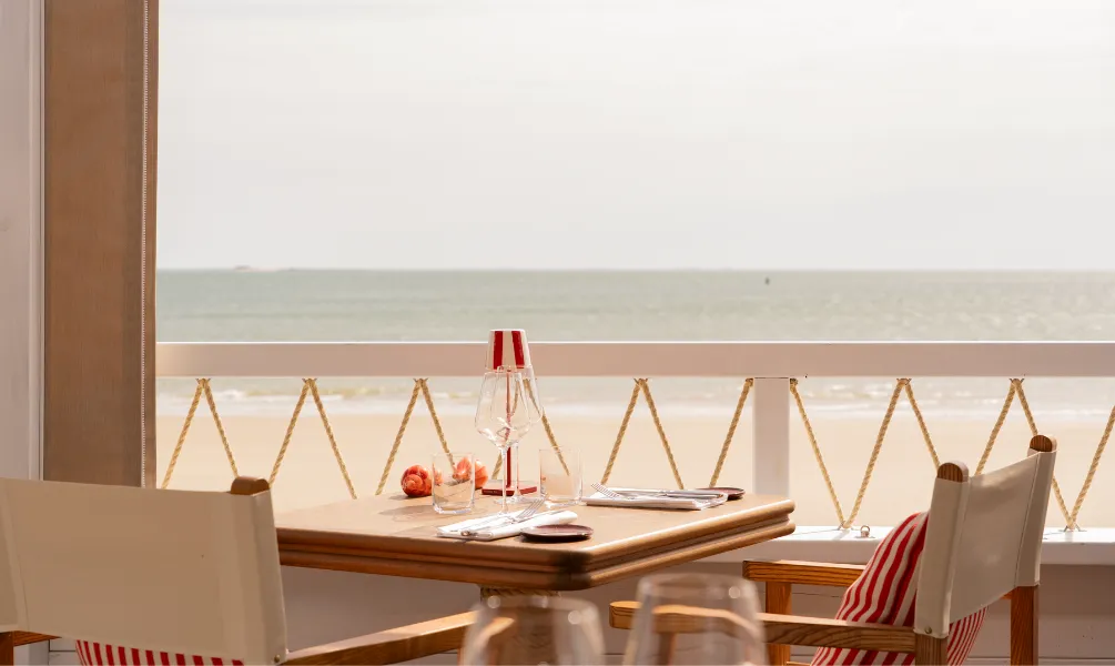 Terrasse de restaurant face à l'océan avec chaises de metteur en scène en bois et toile blanche, coussins rayés rouge et blanc et balustrade en corde nautique.