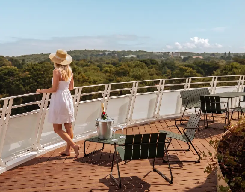 Une bouteille de Champagne avec deux coupes posées sur la table. Une femme observe le paysage depuis une grande terrasse.