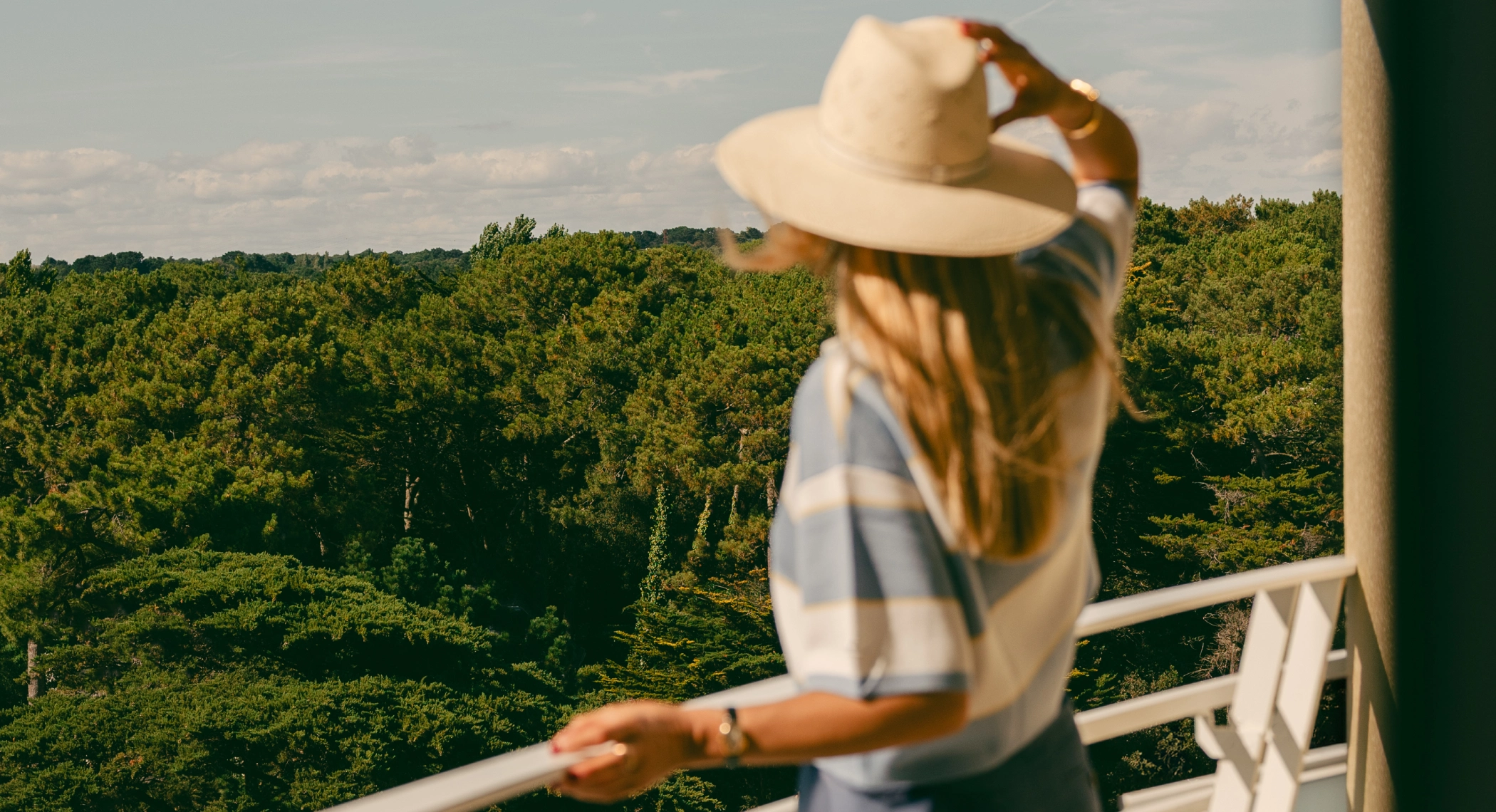 Femme tenant un chapeau et admirant la vue sur la canopée depuis un balcon.