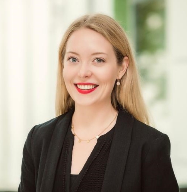 Porträt einer lächelnden Frau mit langen blondem Haar, rotem Lippenstift, schwarzem Blazer und Perlenohrring vor hellem Hintergrund.