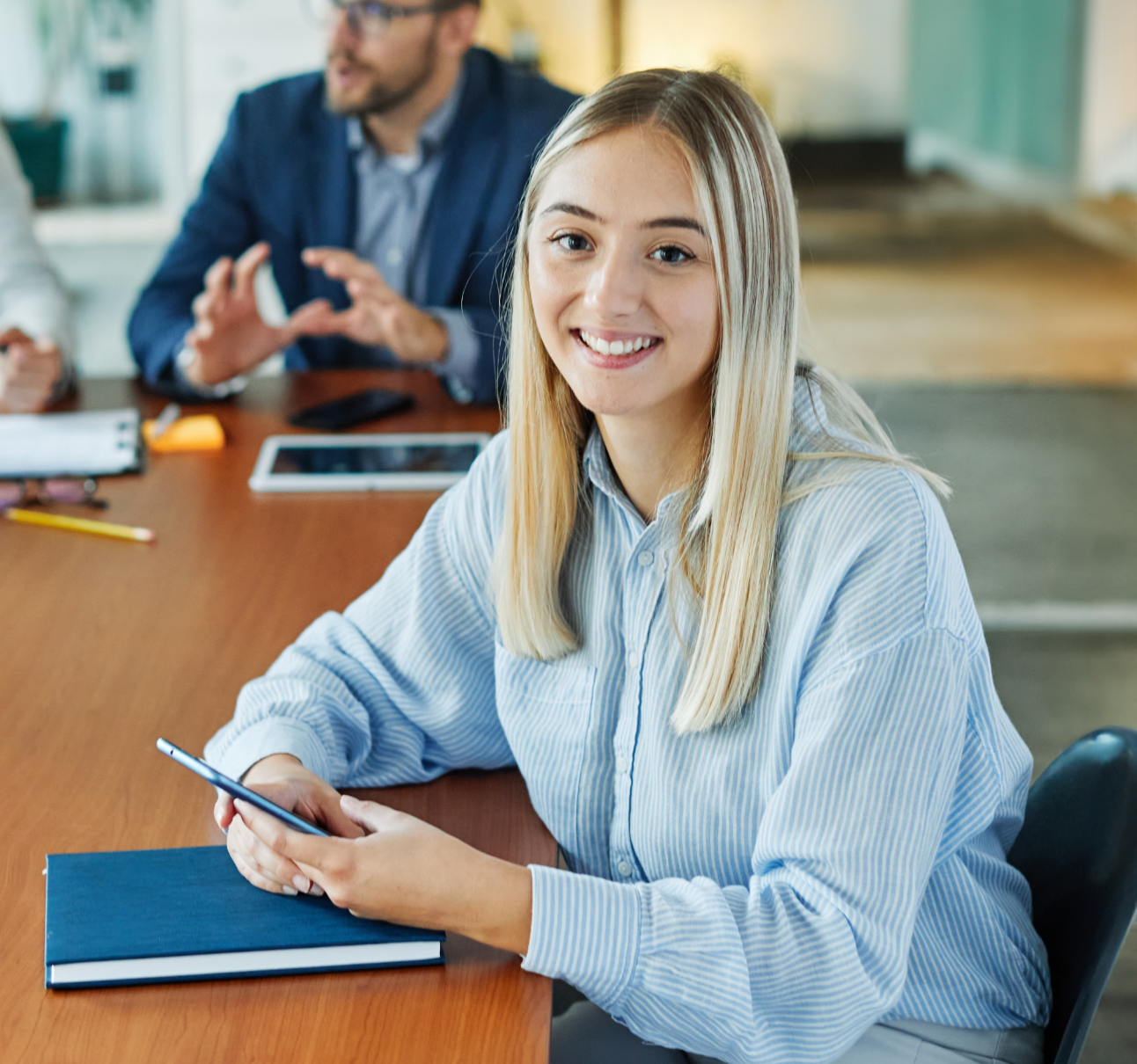 Lächelnde junge Frau in einem hellblau gestreiften Hemd sitzt an einem Besprechungstisch und hält ein Smartphone.