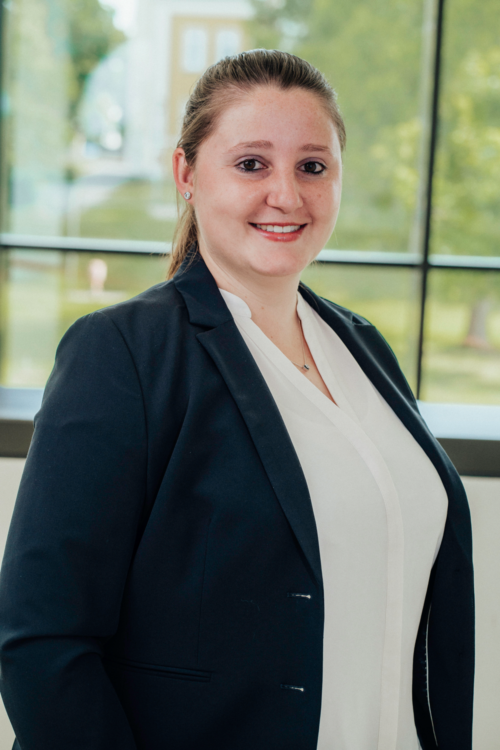 Lächelnde Frau mit zusammengebundenem Haar trägt dunkelblauen Blazer und weiße Bluse vor einem Fenster mit Blick ins Grüne.