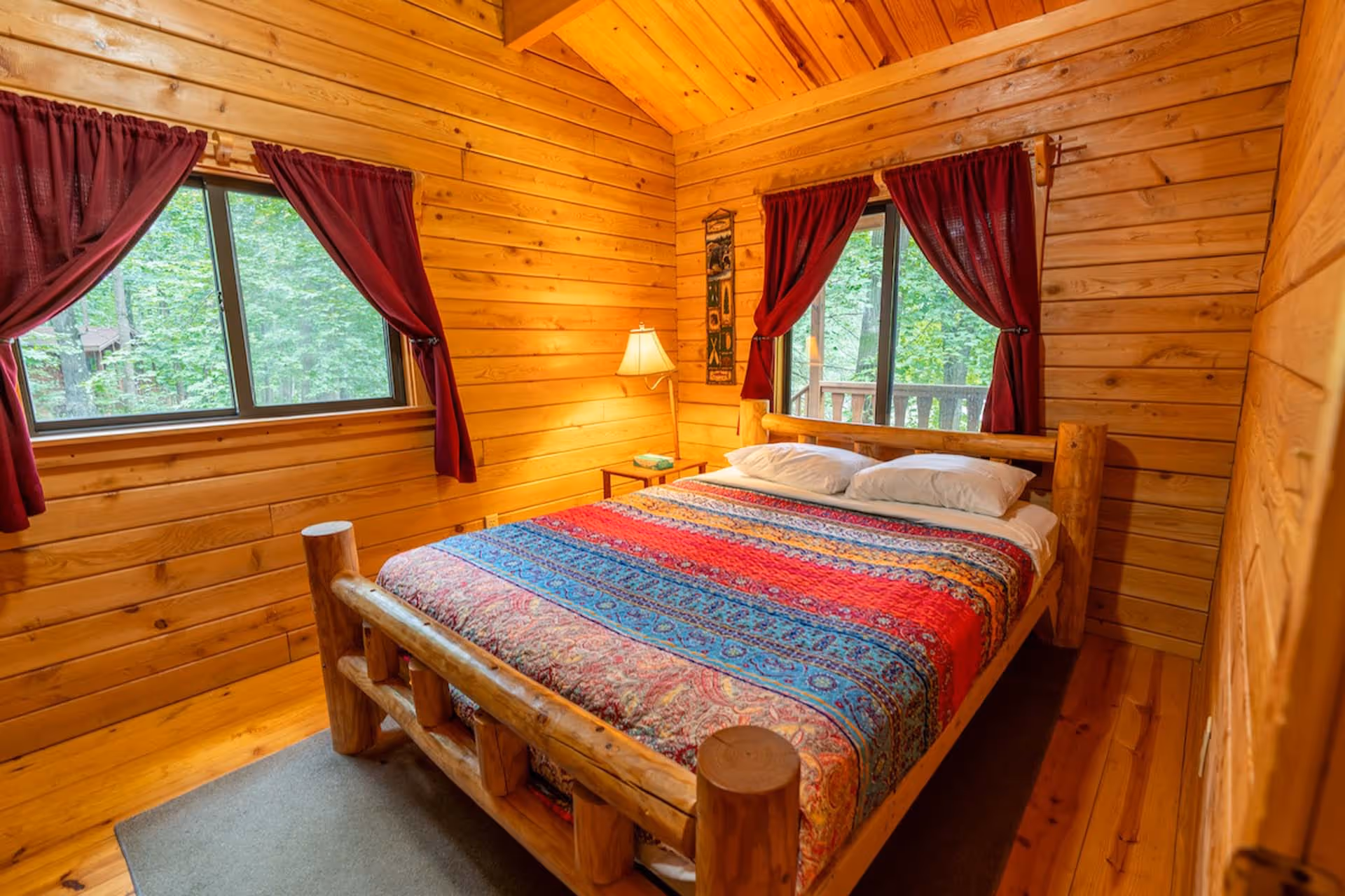 Cozy wooden cabin bedroom with a rustic log bed, colorful patterned bedspread, red curtains, and two windows showing green forest outside.
