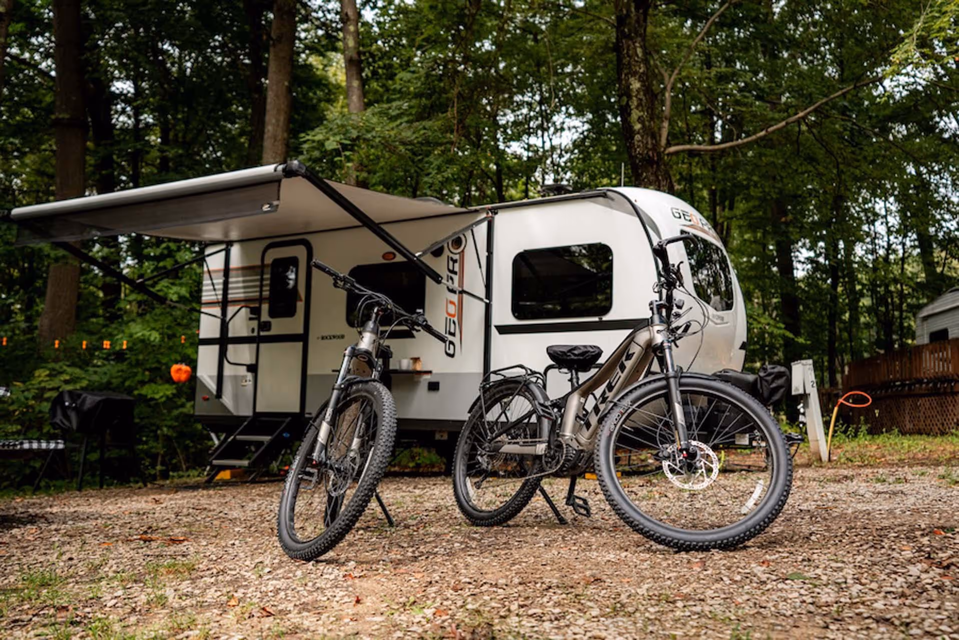 Two mountain bikes parked in front of a white RV trailer with an open awning in a wooded campsite.