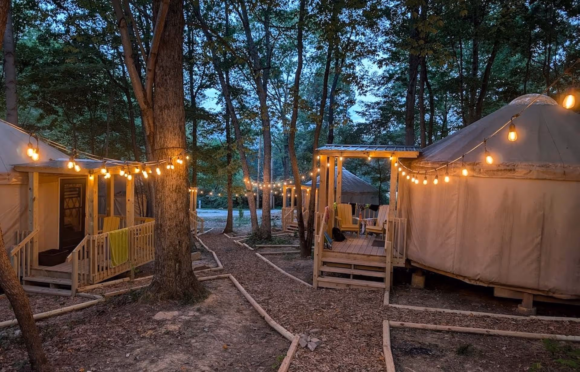 Glamping yurts in a wooded area at dusk with string lights illuminating wooden porches and pathways.