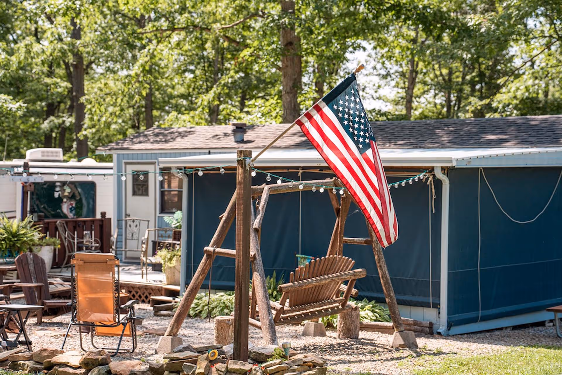Wooden porch swing with American flag attached, surrounded by outdoor chairs and string lights in front of a blue mobile home.