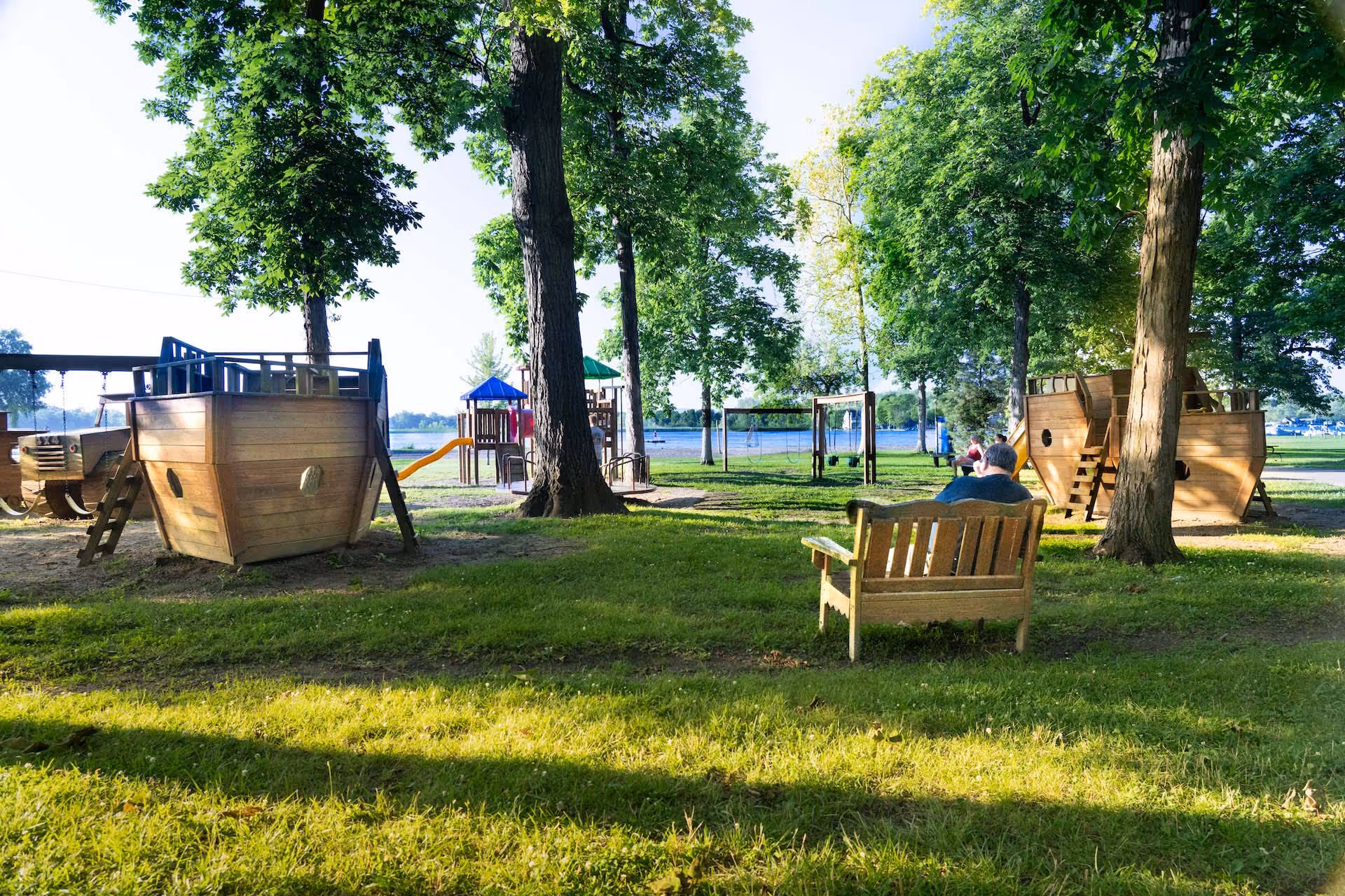 Park playground with wooden ship-shaped structures, swings, slide, and people sitting on benches under green trees near a lake.
