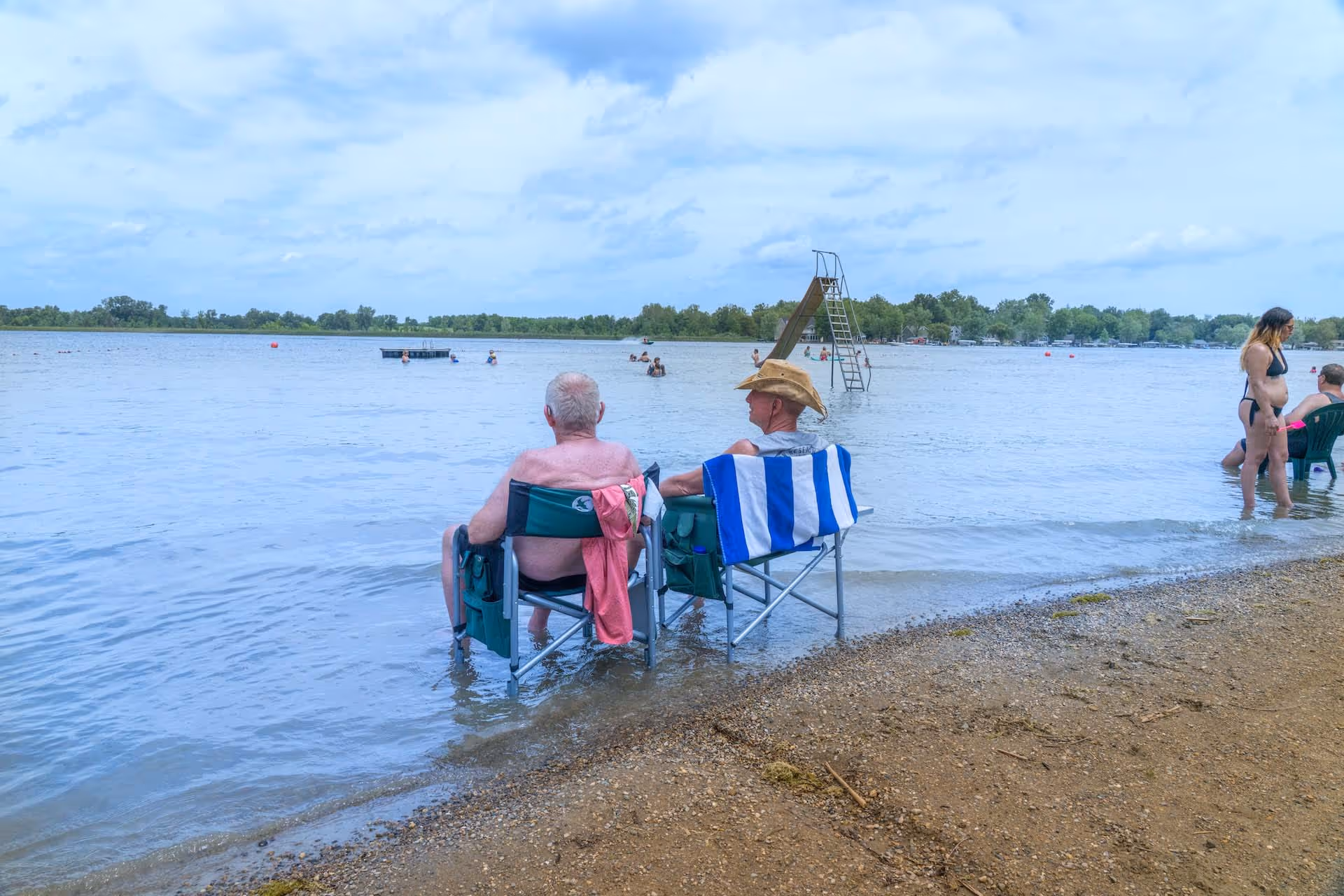 Two men sitting on chairs partially submerged in water at a lake beach with other people swimming and relaxing nearby under a cloudy sky.