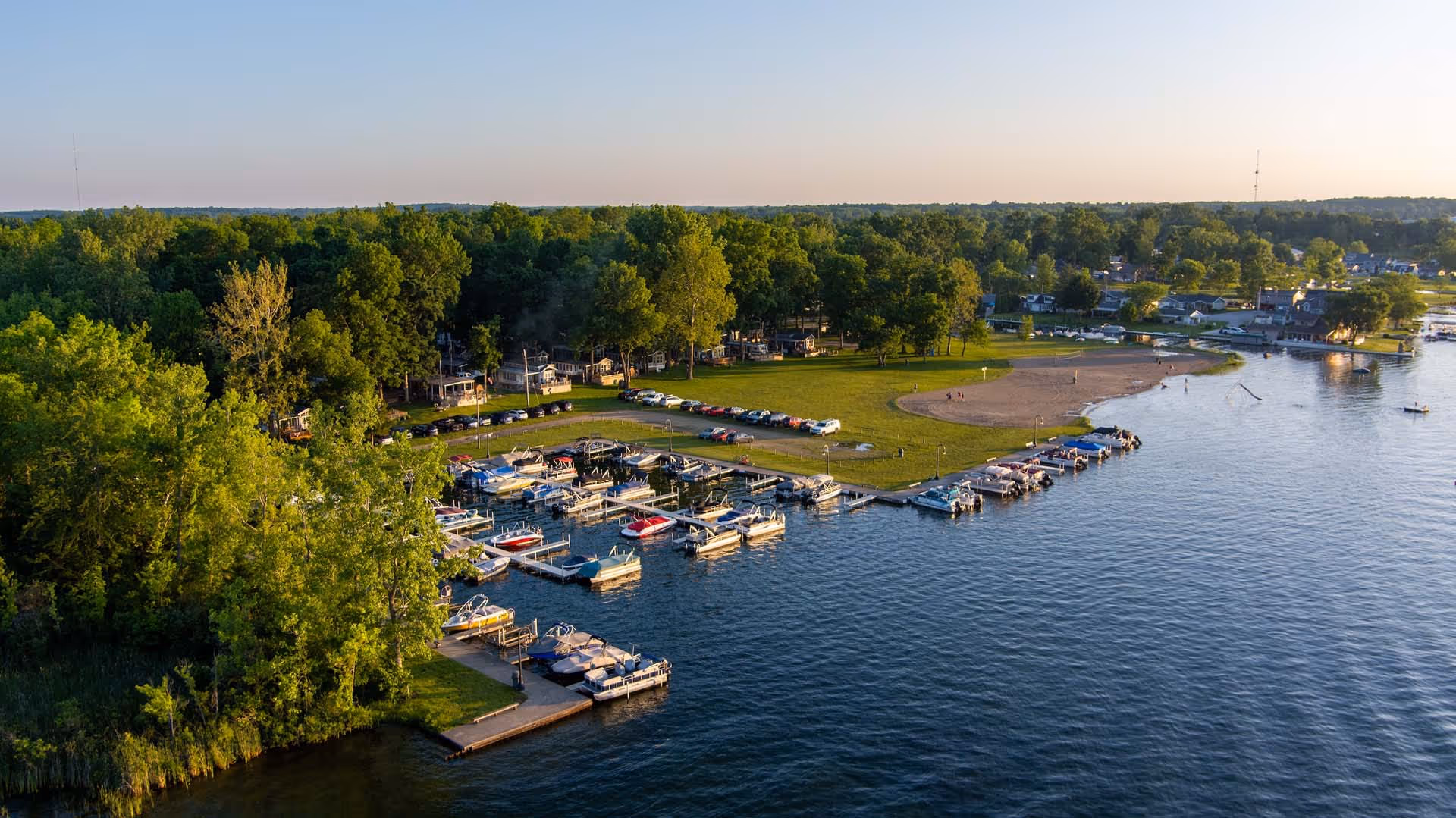 Aerial view of a marina with numerous boats docked, adjacent to a green parkland with parked cars and trees, beside a calm lake.