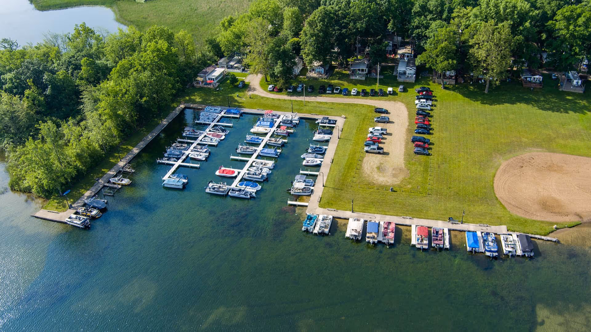 Aerial view of a marina with multiple boats docked, surrounded by green trees and parked cars on grassy land.